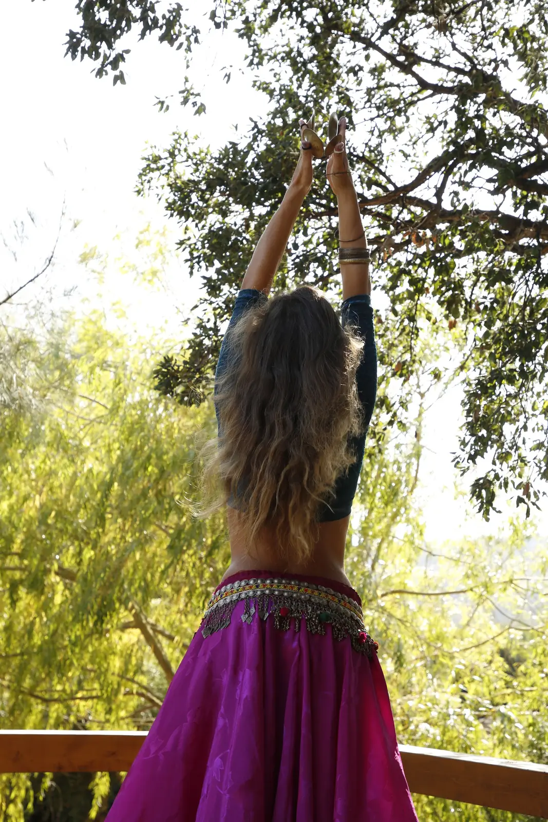 Ana seen from behind, arms raised with finger cymbals and long wavy hair, beneath a tree canopy