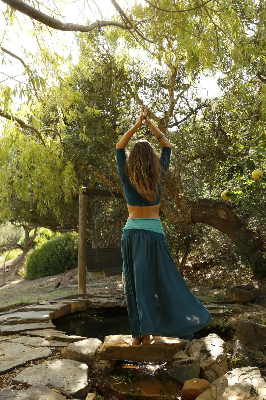 Ana seen from behind, standing on a stone by a small pond with her hands joined above her head, under the trees