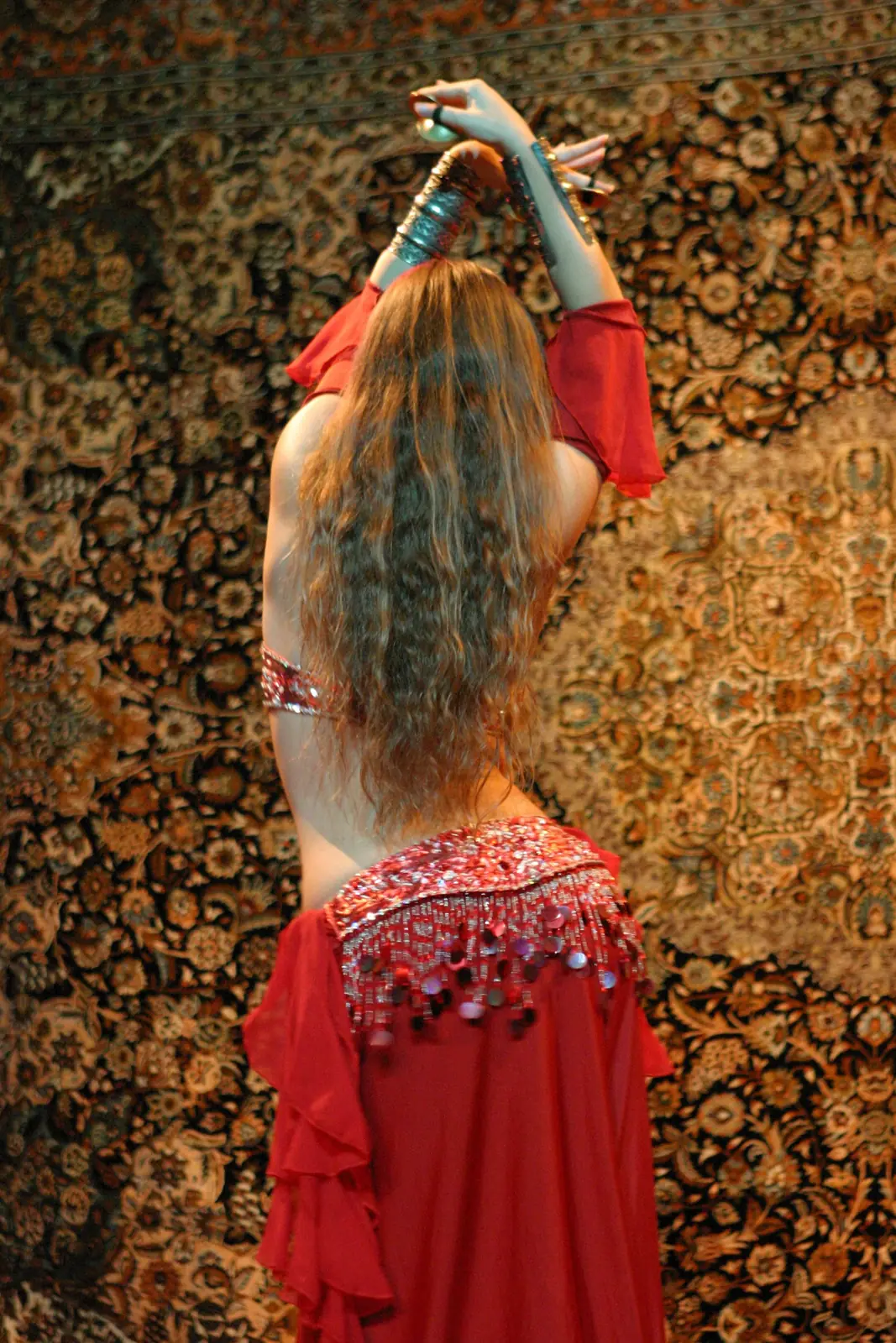 Ana seen from behind, with long wavy hair and a red belly dance costume, arms raised before an oriental carpet