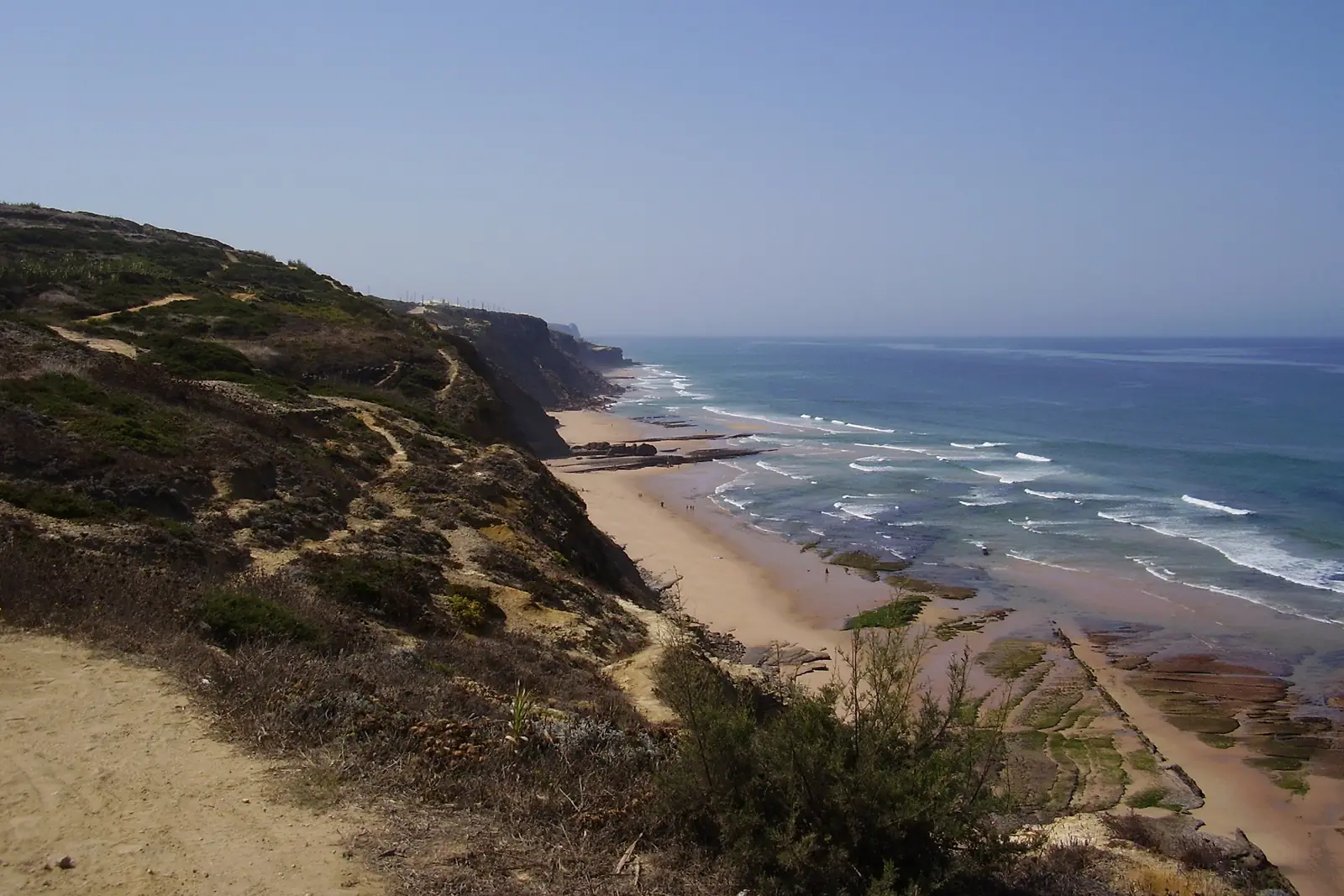 Scrub-covered cliffs and a dirt path above a long sandy beach and the sea on a clear day